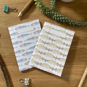Puede incluir: Dos cuadernos pequeños con fondo blanco y un patrón repetitivo de peces. Un cuaderno tiene un patrón de peces azules y el otro tiene un patrón de peces amarillos.