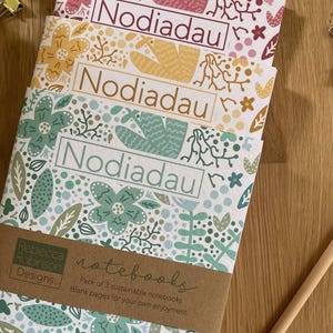 Puede incluir: Un paquete de tres cuadernos con diseños florales en rosa, amarillo y verde azulado. Cada cuaderno presenta la palabra "Nodiadau" y está sujeto con una banda de papel marrón que dice "notebooks". También se ven un lápiz y un clip.
