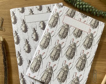 May include: Two notebooks with a white background and a repeating pattern of black and brown beetles. The notebooks have a blank white label on the front cover.