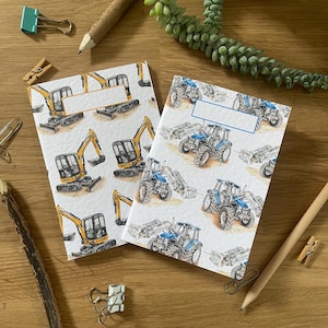 Puede incluir: Dos cuadernos con cubiertas blancas con ilustraciones de vehículos de construcción. Uno tiene excavadoras amarillas y el otro tractores azules. Los cuadernos están sobre una superficie de madera con artículos de papelería.