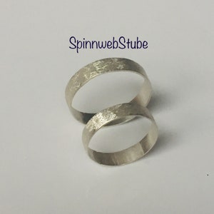 Puede incluir: Dos anillos de plata con superficie texturizada se muestran sobre un fondo blanco. Los anillos están uno al lado del otro, con el texto "SpinnwebStube" en escritura cursiva azul en la esquina superior izquierda. Los anillos parecen ser alianzas de boda.