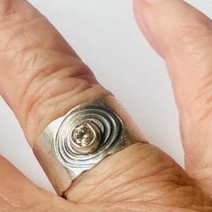 May include: A wide silver band ring featuring a central circular setting with a small, light-coloured gemstone. The metal surface around the stone is textured with concentric, etched circular lines that create a ripple effect across the band.
