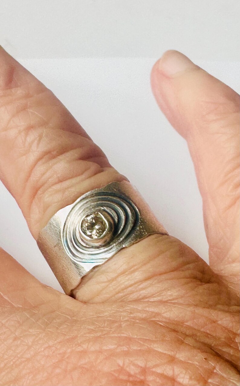 May include: A wide silver band ring featuring a central circular setting with a small, light-coloured gemstone. The metal surface around the stone is textured with concentric, etched circular lines that create a ripple effect across the band.