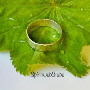 Könnte beinhalten: Ein gehammerter Goldring liegt auf einem leuchtend grünen Blatt mit Wassertropfen. Der Ring hat eine strukturierte Oberfläche und ein schlichtes, elegantes Design. Der Text "SpinnwebStube" ist unten im Bild zu sehen.