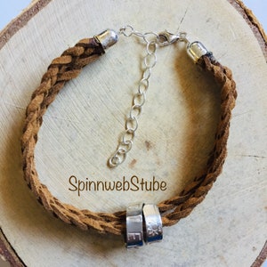 May include: A brown leather braided bracelet with a silver clasp and two silver rings with engraved lettering. The bracelet is on a wooden surface. The text on the bracelet reads "SpinnwebStube".