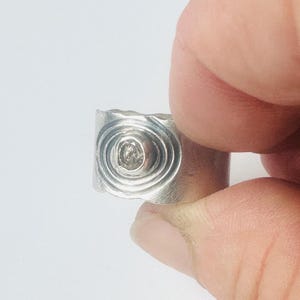 May include: A wide silver band ring featuring a central circular setting with a small, clear, faceted stone. The surface of the ring is decorated with concentric etched circles surrounding the stone, creating a textured, metallic appearance.