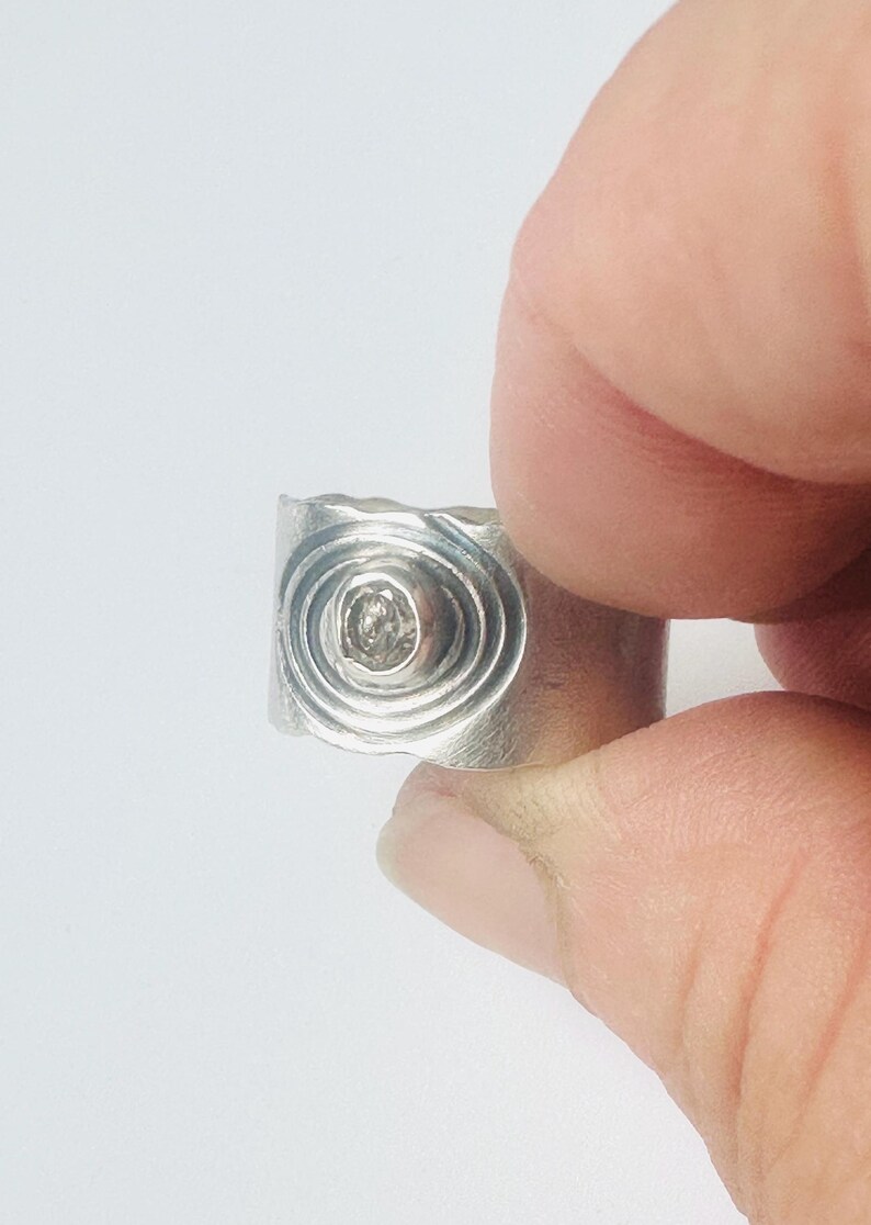May include: A wide silver band ring featuring a central circular setting with a small, clear, faceted stone. The surface of the ring is decorated with concentric etched circles surrounding the stone, creating a textured, metallic appearance.