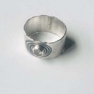 May include: A wide silver band ring with a textured, uneven top edge. The ring features a central circular setting containing a small, clear stone, surrounded by dark, etched concentric circles that create a target-like pattern on the metal surface.