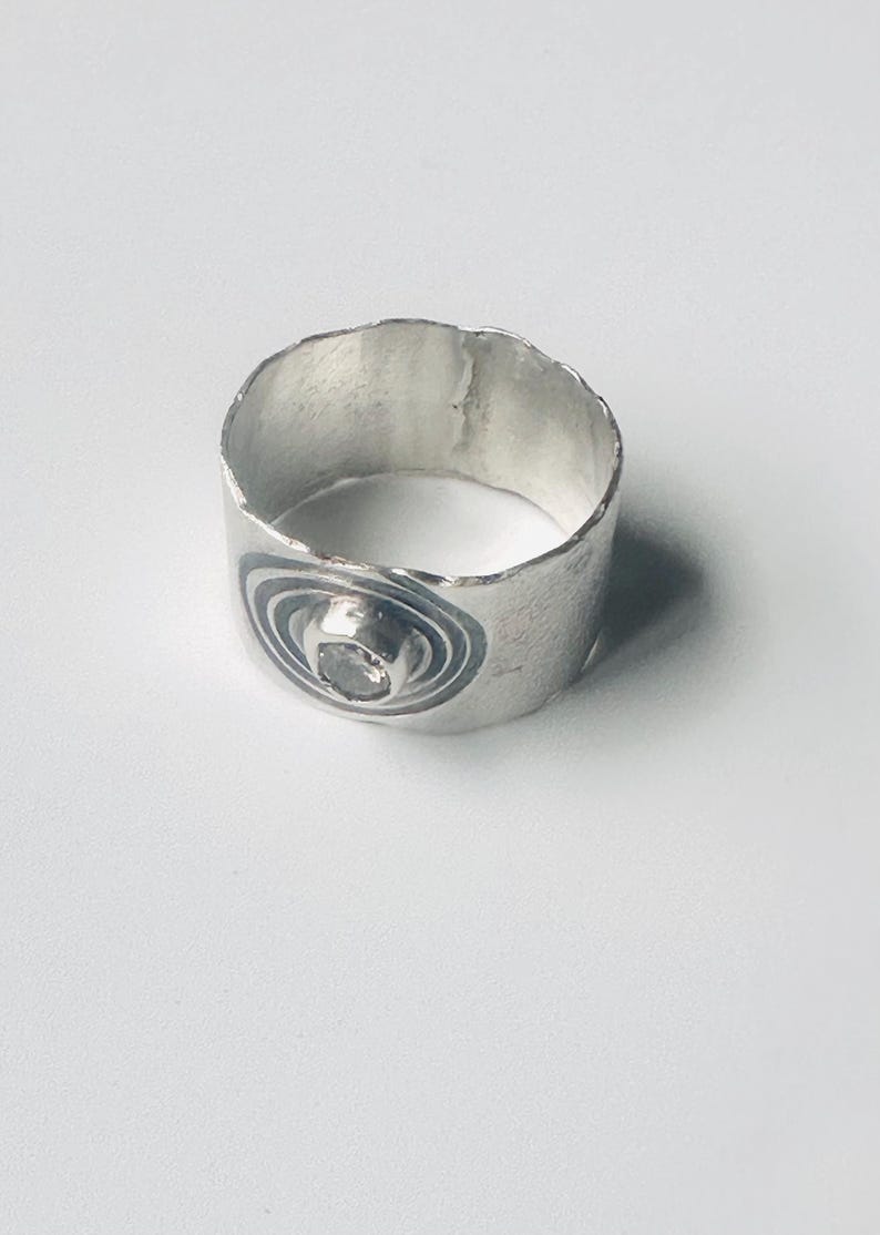 May include: A wide silver band ring with a textured, uneven top edge. The ring features a central circular setting containing a small, clear stone, surrounded by dark, etched concentric circles that create a target-like pattern on the metal surface.