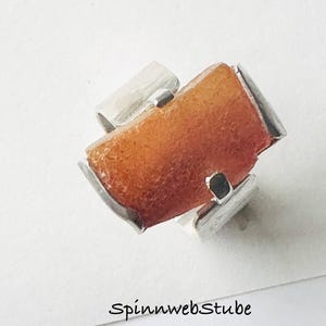 Könnte beinhalten: Ein moderner Ring mit einem rechteckigen, orange-braunen Stein, der in einem silberfarbenen Metallband gefasst ist. Der Ring hat ein einzigartiges Design mit geometrischen Akzenten. Der Text "SpinnwebStube" ist unten im Bild zu sehen.