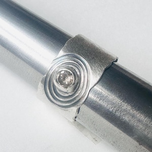 May include: A silver ring featuring a textured, wide band with a central circular motif. The centre of the motif contains a single, small, round, light-coloured gemstone set in a raised bezel. The ring is displayed on a metal mandrel.