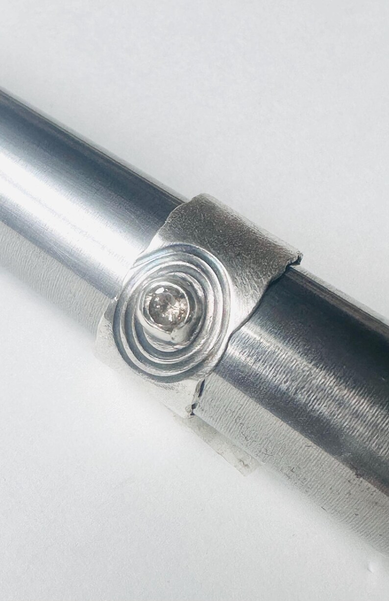 May include: A silver ring featuring a textured, wide band with a central circular motif. The centre of the motif contains a single, small, round, light-coloured gemstone set in a raised bezel. The ring is displayed on a metal mandrel.