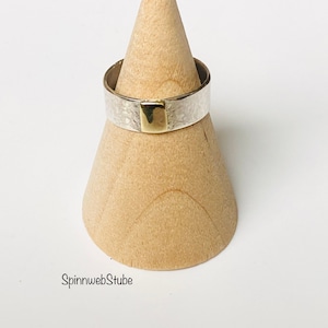 Puede incluir: Un anillo de plata con textura martillada y un acento cuadrado dorado. El anillo se exhibe sobre un soporte de madera en forma de cono sobre un fondo blanco. El diseño del anillo es simple y moderno.