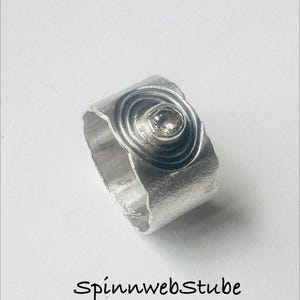 Könnte beinhalten: Ein breiter Silberring mit einer texturierten, matten Oberfläche, der ein zentrales kreisförmiges Motiv mit konzentrischen Ringen und einem kleinen, klaren Edelstein in der Mitte aufweist. Der Text Spinnwebstube ist unter dem Ring schwarz gedruckt.