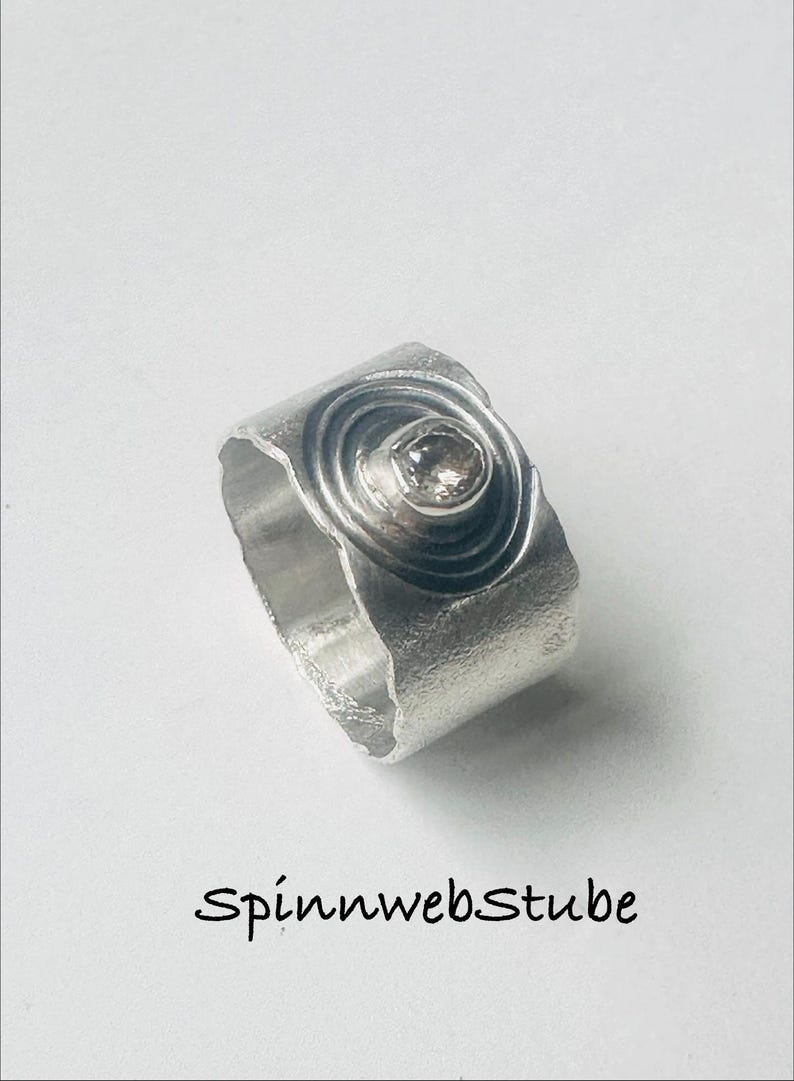May include: A wide silver ring with a textured, matte finish featuring a central circular motif with concentric rings and a single small, clear gemstone set in the middle. The text Spinnwebstube is printed in black below the ring.