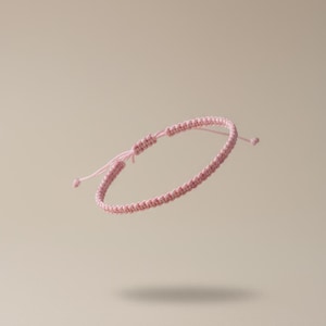 Könnte beinhalten: Ein hellrosa Makramee-Armband mit verstellbarem Schiebeknotenverschluss. Das Armband besteht aus gewebtem Kordel und hat ein schlichtes, minimalistisches Design. Das Armband wird vor einem neutralen Hintergrund gezeigt.