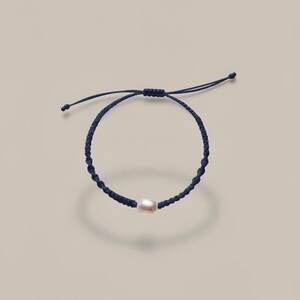 May include: A navy blue macramé bracelet featuring a single, off-white pearl bead. The bracelet is adjustable with sliding knots and is set against a neutral backdrop. A simple and elegant piece of jewellery.