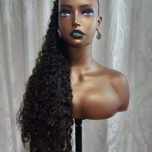 May include: A black curly hair extension on a mannequin head. The hair is long and reaches the mannequin's waist. The mannequin is wearing a black headband.