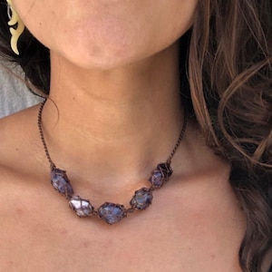 Puede incluir: Un collar hecho a mano con piedras de amatista en bruto, envueltas en un alambre tejido marrón oscuro. El collar tiene una cadena delicada y muestra los tonos naturales púrpura y blanco de las piedras preciosas. Una joya única.
