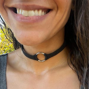 May include: Black macrame choker necklace with a silver-colored ring in the center. The choker is worn around the neck. The background is blurred, with green foliage visible.