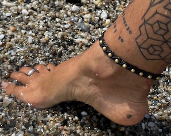 May include: Black macramé anklet with gold-coloured beads. The anklet is worn on an ankle with a tattoo. The foot is wet, and the background is a beach with small pebbles.