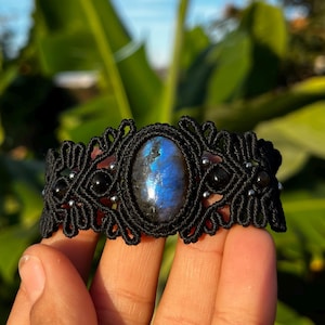 May include: Black macramé bracelet with a central oval labradorite stone, displaying blue and grey flashes. The bracelet incorporates black onyx beads and silver-toned details. The background shows green leaves and a blue sky.