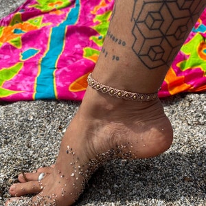 Könnte beinhalten: Nahaufnahme eines Knöchels, der ein hellbraunes Fußkettchen mit goldenen Perlen trägt. Das Armband ist aus geflochtenem Band gefertigt und hat ein schlichtes Design. Die Person steht auf einem Strand mit Sand und Kieselsteinen.