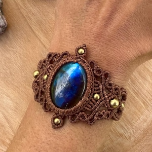 May include: A brown macrame bracelet with a large, oval, blue gemstone at the center. The bracelet is adorned with small gold-colored beads. The gemstone has a blue and green iridescent quality. The bracelet is on a wooden surface.