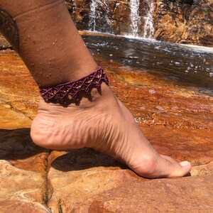 May include: A close-up of a burgundy ankle bracelet with gold beads. The bracelet is made of a delicate, intricate design. The bracelet is worn on a person's ankle, with the foot resting on a rock.