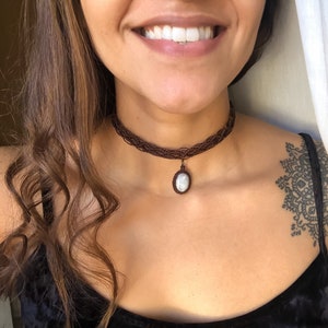 K&ouml;nnte beinhalten: Ein braunes Makramee-Choker-Halskette mit einem ovalen Anh&auml;nger. Der Anh&auml;nger hat einen hellfarbigen Stein. Die Halskette wird um den Hals getragen. Der Choker besteht aus gewebten braunen F&auml;den.