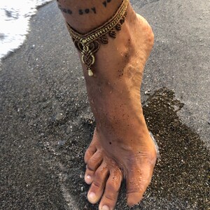 Puede incluir: Una tobillera de macram&eacute; dorada con un colgante decorativo. La tobillera se lleva en un pie al borde del oc&eacute;ano. El pie est&aacute; parcialmente sumergido en el agua, con arena oscura visible.