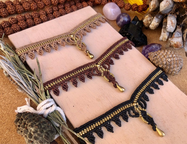 May include: Three macrame headbands in tan, brown, and black, each with gold-colored accents and a teardrop pendant. The headbands are displayed on a light brown surface, with natural elements in the background.