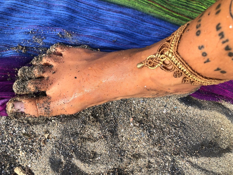Puede incluir: Una tobillera de macram&eacute; dorada con un dise&ntilde;o decorativo, con un peque&ntilde;o dije colgante. La tobillera se lleva en un pie cubierto de arena, con una tela colorida en el fondo. La imagen est&aacute; tomada al aire libre.