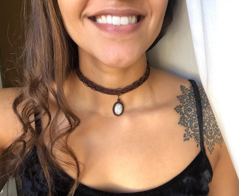 K&ouml;nnte beinhalten: Ein braunes Makramee-Choker-Halskette mit einem ovalen Anh&auml;nger. Der Anh&auml;nger hat einen hellfarbigen Stein. Die Halskette wird um den Hals getragen. Der Choker besteht aus gewebten braunen F&auml;den.