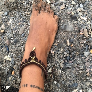 Puede incluir: Una tobillera de macram&eacute; marr&oacute;n y dorada con un dise&ntilde;o de corona. La tobillera se lleva en un tobillo cubierto de arena, con un tatuaje visible debajo. El fondo es una playa con peque&ntilde;as rocas y arena.