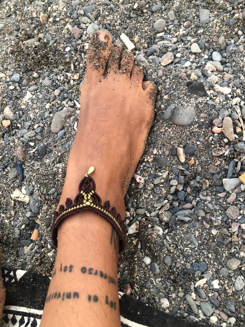 Puede incluir: Una tobillera de macram&eacute; marr&oacute;n y dorada con un dise&ntilde;o de corona. La tobillera se lleva en un tobillo cubierto de arena, con un tatuaje visible debajo. El fondo es una playa con peque&ntilde;as rocas y arena.