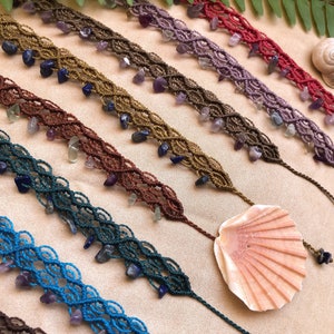 Può includere: Braccialetti macramè fatti a mano in vari colori, adornati con perline di pietre preziose. I braccialetti sono esposti su una superficie neutra, con una conchiglia e fronde di felce sullo sfondo. Ogni braccialetto misura circa 18 cm di lunghezza.