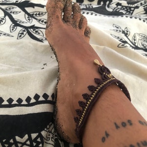Puede incluir: Primer plano de un pie cubierto de arena, adornado con una tobillera marr&oacute;n y dorada con un dise&ntilde;o de hojas. Un tatuaje en el tobillo dice "Yo soy el universo". El pie descansa sobre una tela estampada en blanco y negro.