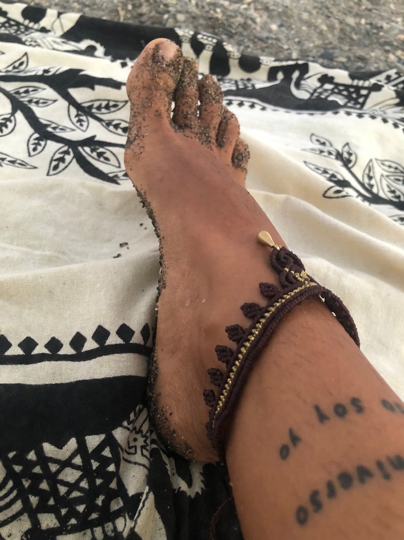 Puede incluir: Primer plano de un pie cubierto de arena, adornado con una tobillera marr&oacute;n y dorada con un dise&ntilde;o de hojas. Un tatuaje en el tobillo dice "Yo soy el universo". El pie descansa sobre una tela estampada en blanco y negro.
