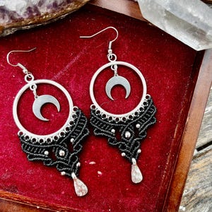May include: A pair of silver-toned hoop earrings with a crescent moon charm and black macrame detailing. The earrings have a teardrop-shaped bead at the bottom. The earrings are displayed on a red velvet surface.