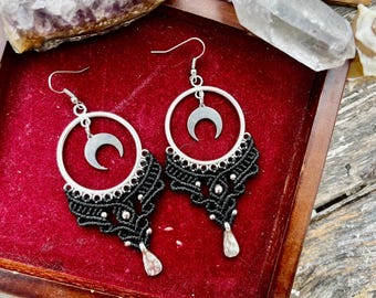 May include: A pair of silver-toned hoop earrings with a crescent moon charm and black macrame detailing. The earrings have a teardrop-shaped bead at the bottom. The earrings are displayed on a red velvet surface.