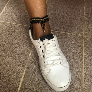 Puede incluir: Una tobillera negra y dorada con detalle de borla se lleva sobre una zapatilla blanca. La tobillera est&aacute; hecha de tela negra con cuentas doradas y una peque&ntilde;a borla dorada. La zapatilla tiene cordones blancos y una suela blanca.