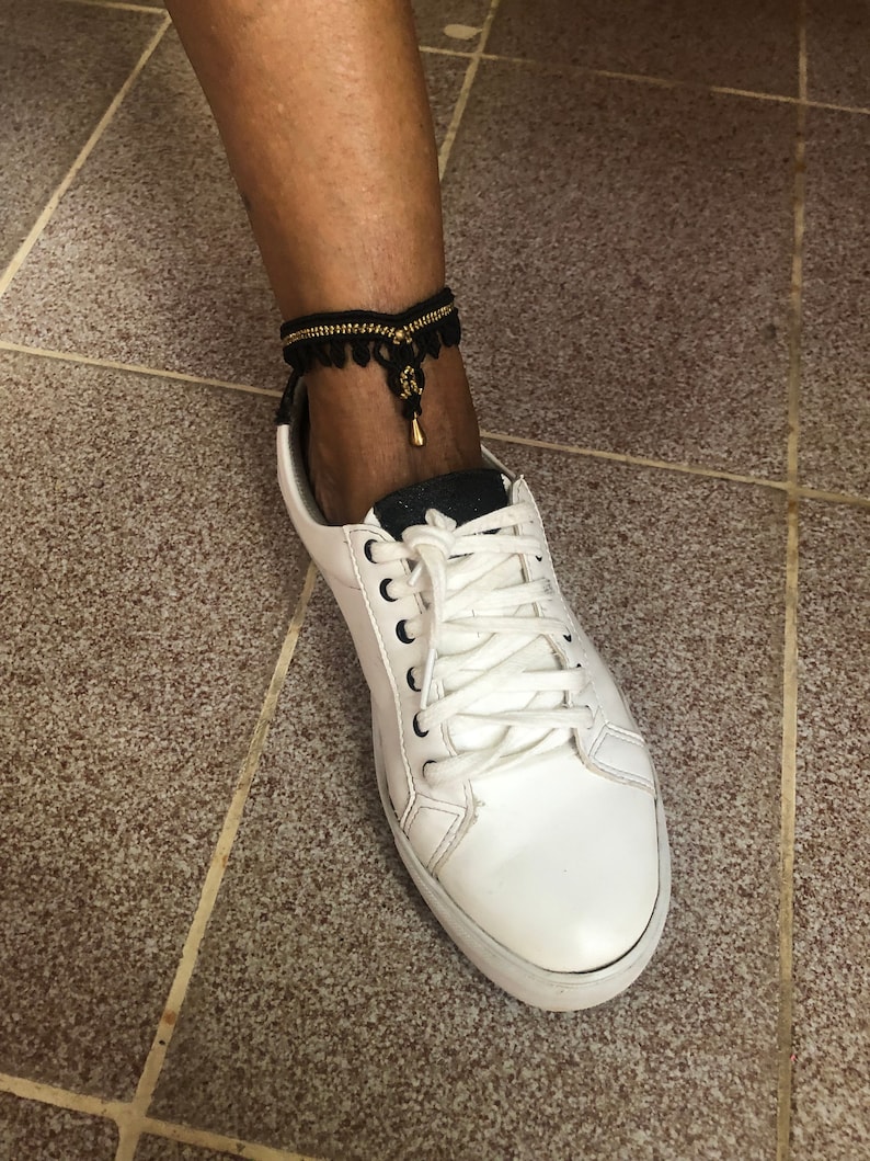 Puede incluir: Una tobillera negra y dorada con detalle de borla se lleva sobre una zapatilla blanca. La tobillera est&aacute; hecha de tela negra con cuentas doradas y una peque&ntilde;a borla dorada. La zapatilla tiene cordones blancos y una suela blanca.