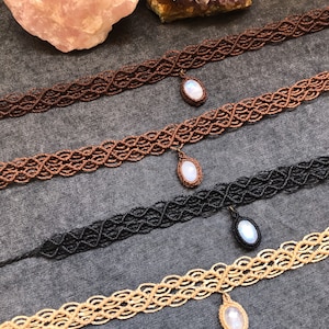 K&ouml;nnte beinhalten: Vier Makramee-Choker in den Farben Braun, Kupfer, Schwarz und Beige. Jeder Choker hat ein gewebtes Muster und einen Anh&auml;nger mit einem wei&szlig;en, ovalen Stein. Die Halsketten werden auf einer grauen Oberfl&auml;che pr&auml;sentiert.