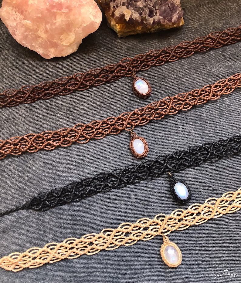 K&ouml;nnte beinhalten: Vier Makramee-Choker in den Farben Braun, Kupfer, Schwarz und Beige. Jeder Choker hat ein gewebtes Muster und einen Anh&auml;nger mit einem wei&szlig;en, ovalen Stein. Die Halsketten werden auf einer grauen Oberfl&auml;che pr&auml;sentiert.