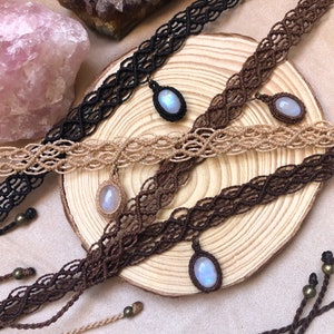 K&ouml;nnte beinhalten: Makramee-Choker in Schwarz, Beige und Braun mit ovalen Edelsteinanh&auml;ngern. Die Halsketten werden auf einer Holzoberfl&auml;che pr&auml;sentiert, mit einem rosafarbenen Kristall und anderen dekorativen Elementen im Hintergrund. Das Makramee ist aufwendig gewebt.