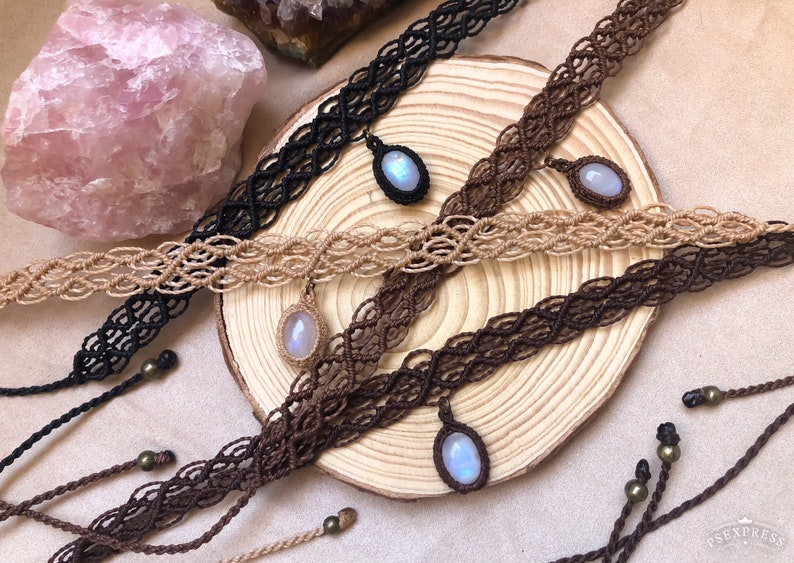 K&ouml;nnte beinhalten: Makramee-Choker in Schwarz, Beige und Braun mit ovalen Edelsteinanh&auml;ngern. Die Halsketten werden auf einer Holzoberfl&auml;che pr&auml;sentiert, mit einem rosafarbenen Kristall und anderen dekorativen Elementen im Hintergrund. Das Makramee ist aufwendig gewebt.