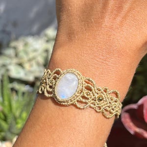 Può includere: Un braccialetto in macramè beige con una pietra di luna ovale al centro. Il braccialetto è realizzato con cordoncino beige con nodi intricati e piccole perline dorate. La pietra di luna presenta un'iridescenza bianca e blu perlata.