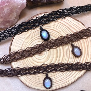 K&ouml;nnte beinhalten: Macram&eacute;-Choker in Schwarz und Braun, jeweils mit einem Anh&auml;nger mit einem hellblauen Edelstein. Die Halsketten werden auf einer Holzoberfl&auml;che pr&auml;sentiert, die die komplizierten Kn&uuml;pfmuster und das subtile Leuchten des Edelsteins hervorhebt.