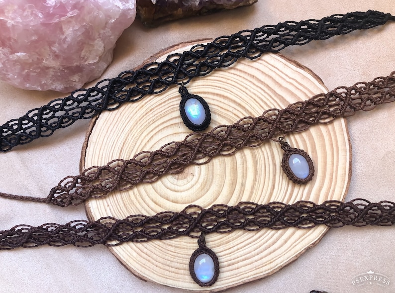 K&ouml;nnte beinhalten: Macram&eacute;-Choker in Schwarz und Braun, jeweils mit einem Anh&auml;nger mit einem hellblauen Edelstein. Die Halsketten werden auf einer Holzoberfl&auml;che pr&auml;sentiert, die die komplizierten Kn&uuml;pfmuster und das subtile Leuchten des Edelsteins hervorhebt.
